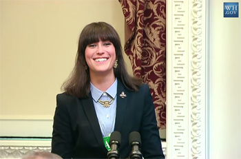 A white woman with dark hair speaks while standing behind a podium